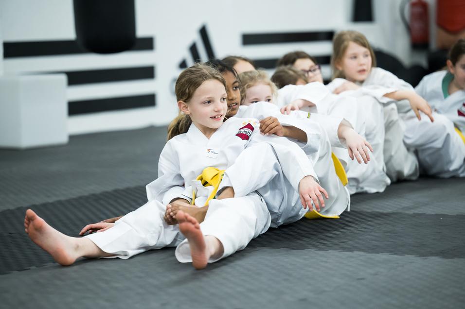 Gruppe von Kindern in weißen Karate-Anzügen sitzt auf einer Matte, einige mit gelben Gürteln, amüsiert und locker.