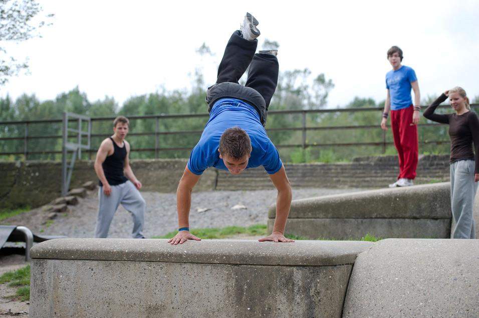 Junger Mann im blauen T-Shirt führt einen Handstand auf einer Betonmauer aus, während andere zuschauen.