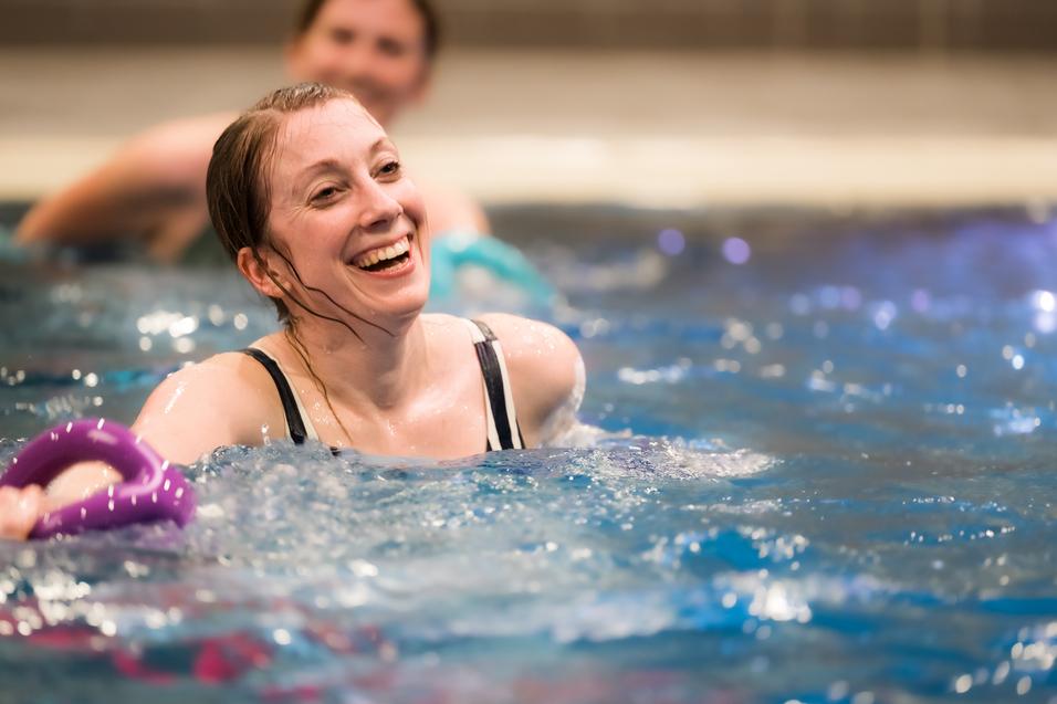 Lächelnde Frau im Wasser hält einen lila Schwimmring, im Hintergrund sind weitere Schwimmer zu sehen.